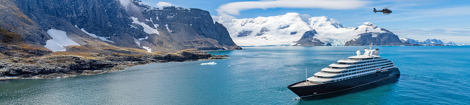 Scenic yacht at sea in Antarctica with a helicopter flying overhead and snow-capped mountains in the backgorund